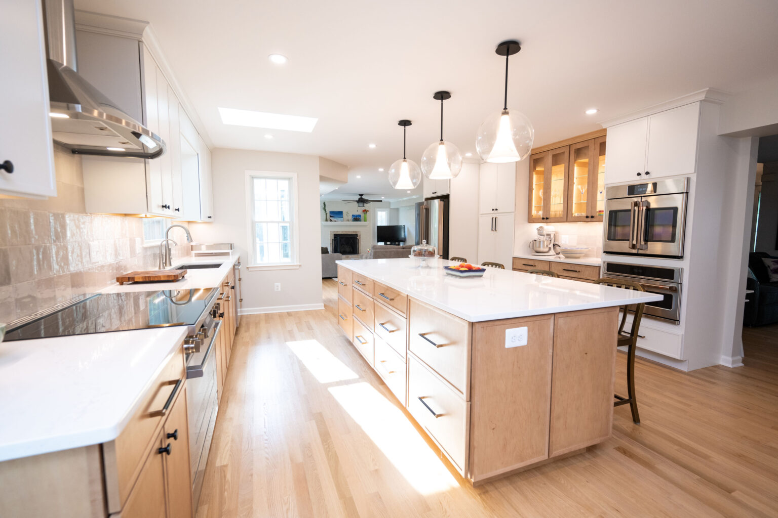 Spacious modern kitchen with light wood floors, white countertops, wooden cabinets, a large central island, and pendant lights. Appliances include a stove, double oven, and microwave.
