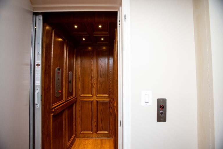 An open elevator with wooden paneling and lit ceiling is visible. A control panel on the wall shows an illuminated button for the second floor.