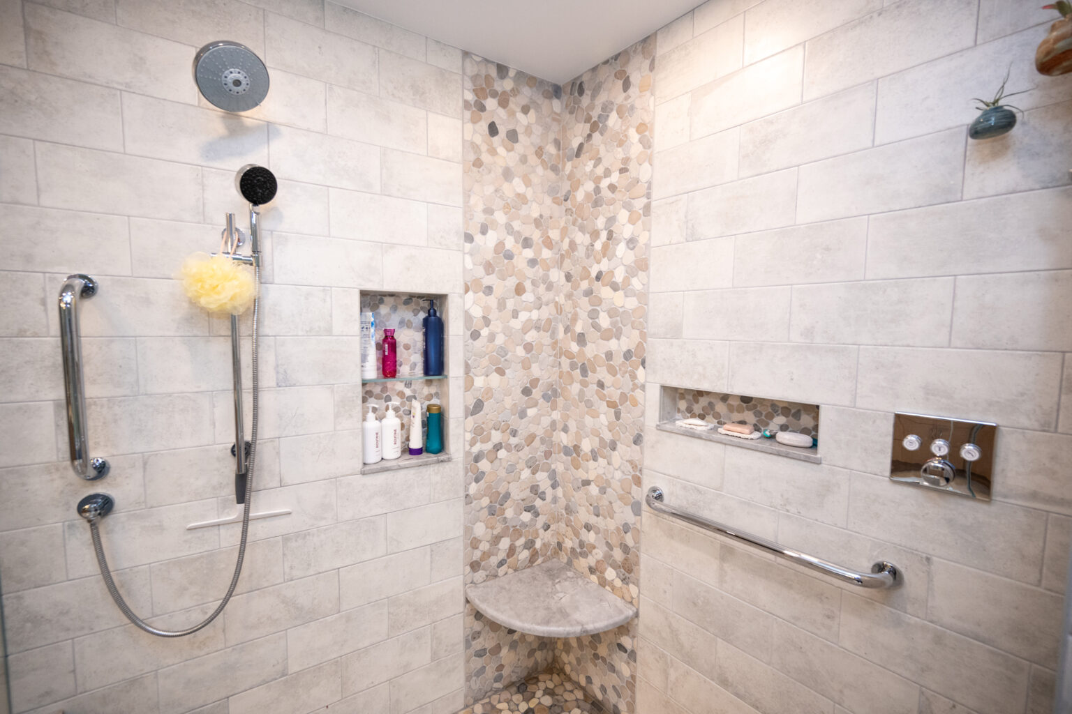 Image by Lesnick Photo Modern shower with built-in shelves holding various bottles, a handheld showerhead, grab bar, corner seat, and pebble-patterned accent tiles on the wall and floor.
