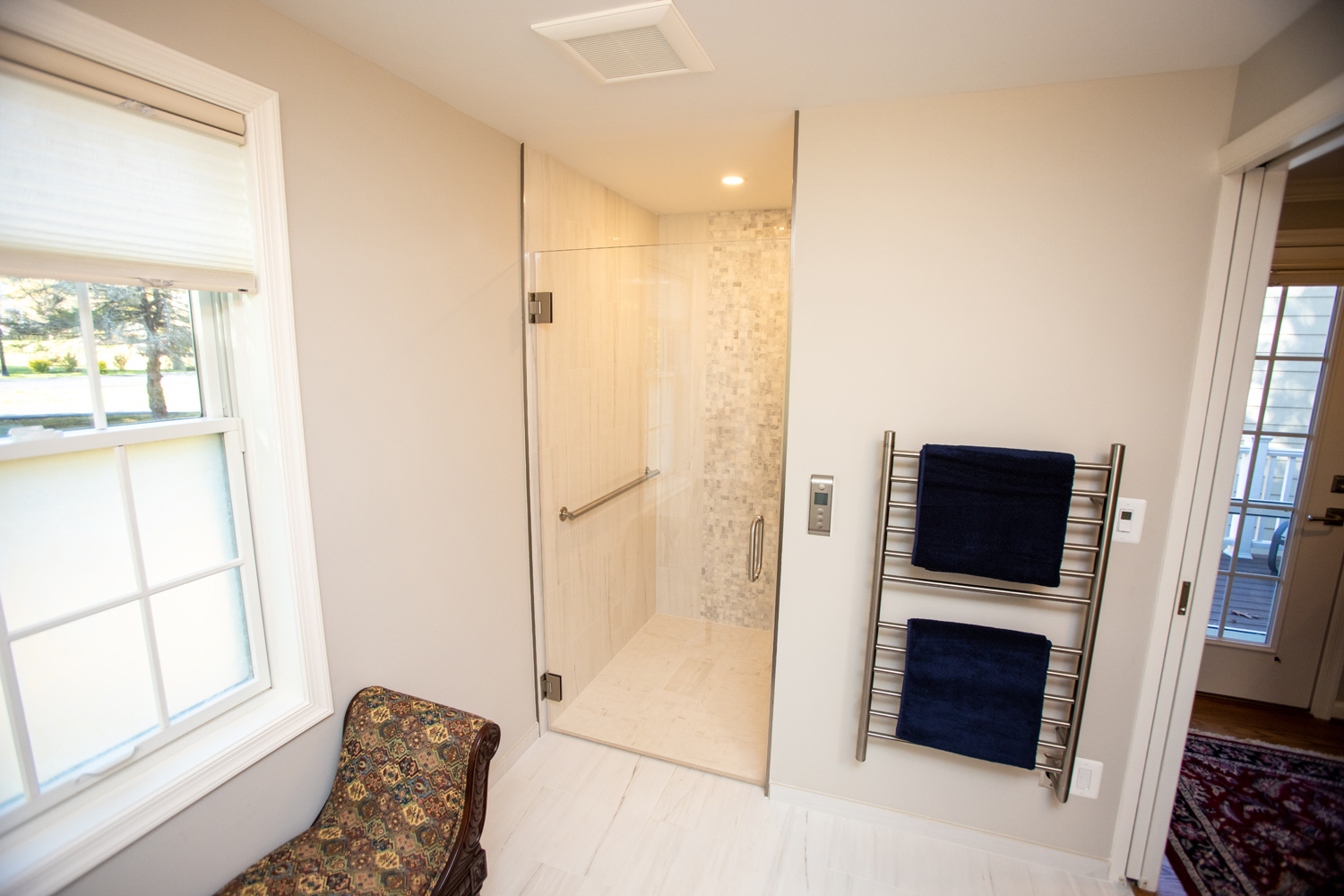Bathroom with a walk-in shower, light beige walls, and a towel rack holding two dark blue towels. A window with a shade is on the left.
