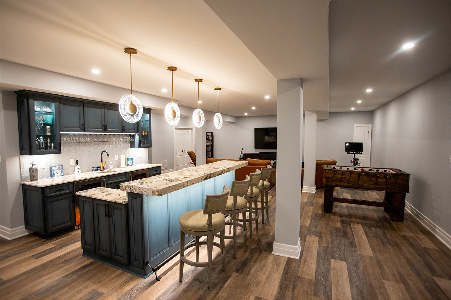 NorthwoodConstruction-Basement-Remodel-Northern-Virginia-couch A spacious basement in Northern Virginia with a bar, tall chairs, a foosball table, leather sofas, and a large TV. This home addition features wooden flooring and modern lighting for ultimate comfort.