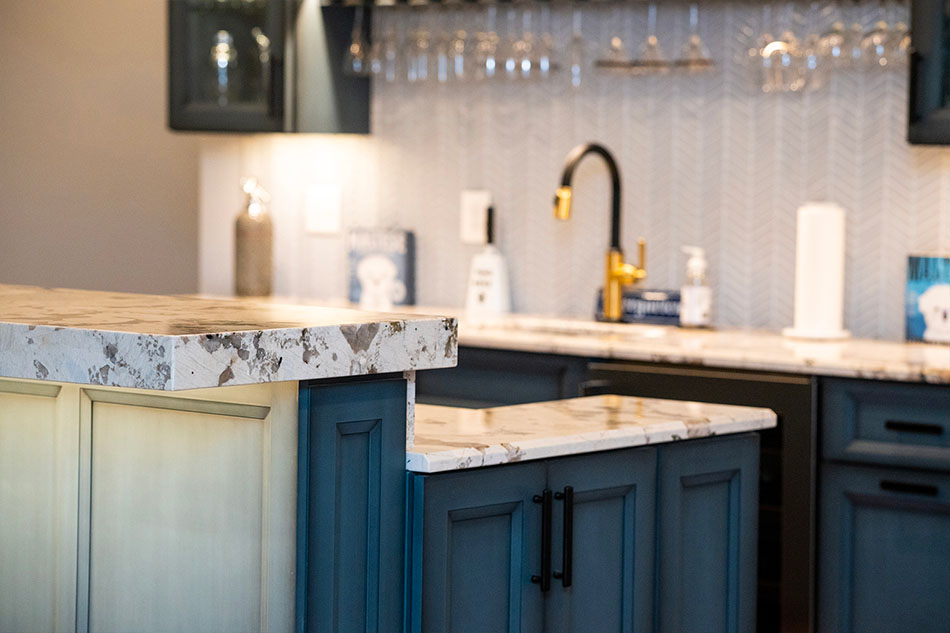NorthwoodConstruction-Basement-Remodel-Northern-Virginia-lighting-bar A modern kitchen with blue cabinets, marble countertops, a black faucet, and glassware hanging above the sink offers a perfect blend of style and functionality—a prime example of home additions in Northern Virginia.