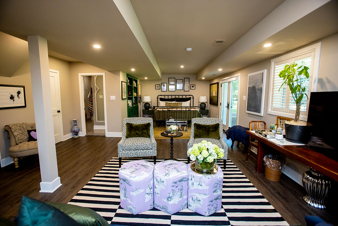 NorthwoodConstruction-Basement-Remodel-Northern-Virginia-open-space A cozy living room with patterned chairs, floral ottomans, a dark wood floor, and a bed in the back. Decor includes framed wall art, a plant, and assorted furniture.