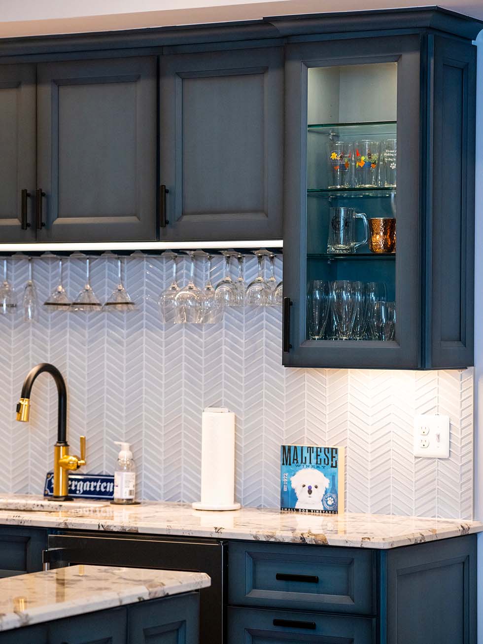 NorthwoodConstruction-Basement-remodel-northern-virginia-sink A chic kitchen in Northern Virginia boasts dark blue cabinets, a gold faucet, and marble countertops. The herringbone tile backsplash adds elegance, while stemware sits on display. A book titled "Maltese" graces the countertop, showcasing thoughtful home additions for a refined touch.