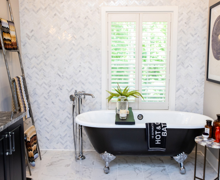 A bathroom with a clawfoot bathtub, marble flooring, a ladder towel rack, and a wall with chevron tiles. The tub has a tray with a candle and a small plant. A window with shutters is behind the tub.