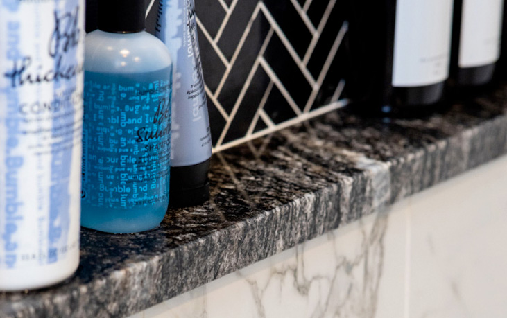 The bathroom shelf is adorned with various shampoo and conditioner bottles, set against a chic black and white herringbone tile backsplash and a sleek marble countertop edge—perfect inspiration for bathroom remodeling in Northern Virginia.