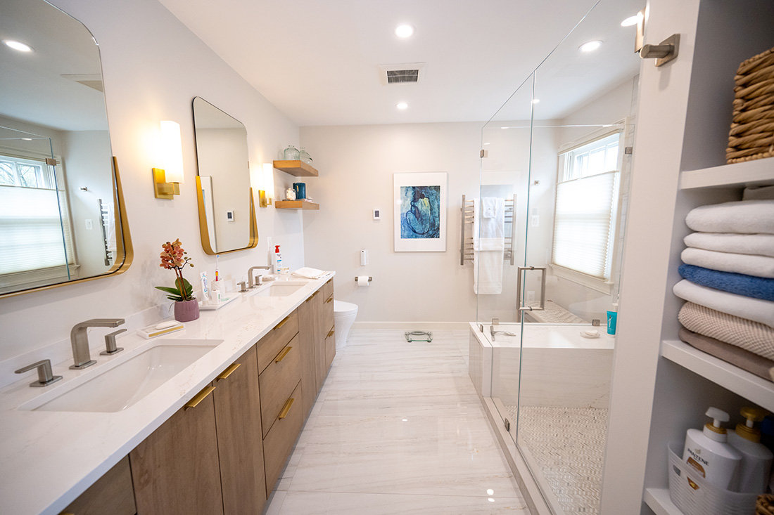 Modern bathroom with double sinks, large mirrors, and gold faucets in a chic Northern Virginia style. A glass-enclosed shower is next to a tub. Shelves hold towels and toiletries. A framed picture hangs above the toilet, showcasing recent bathroom remodeling trends in the area.