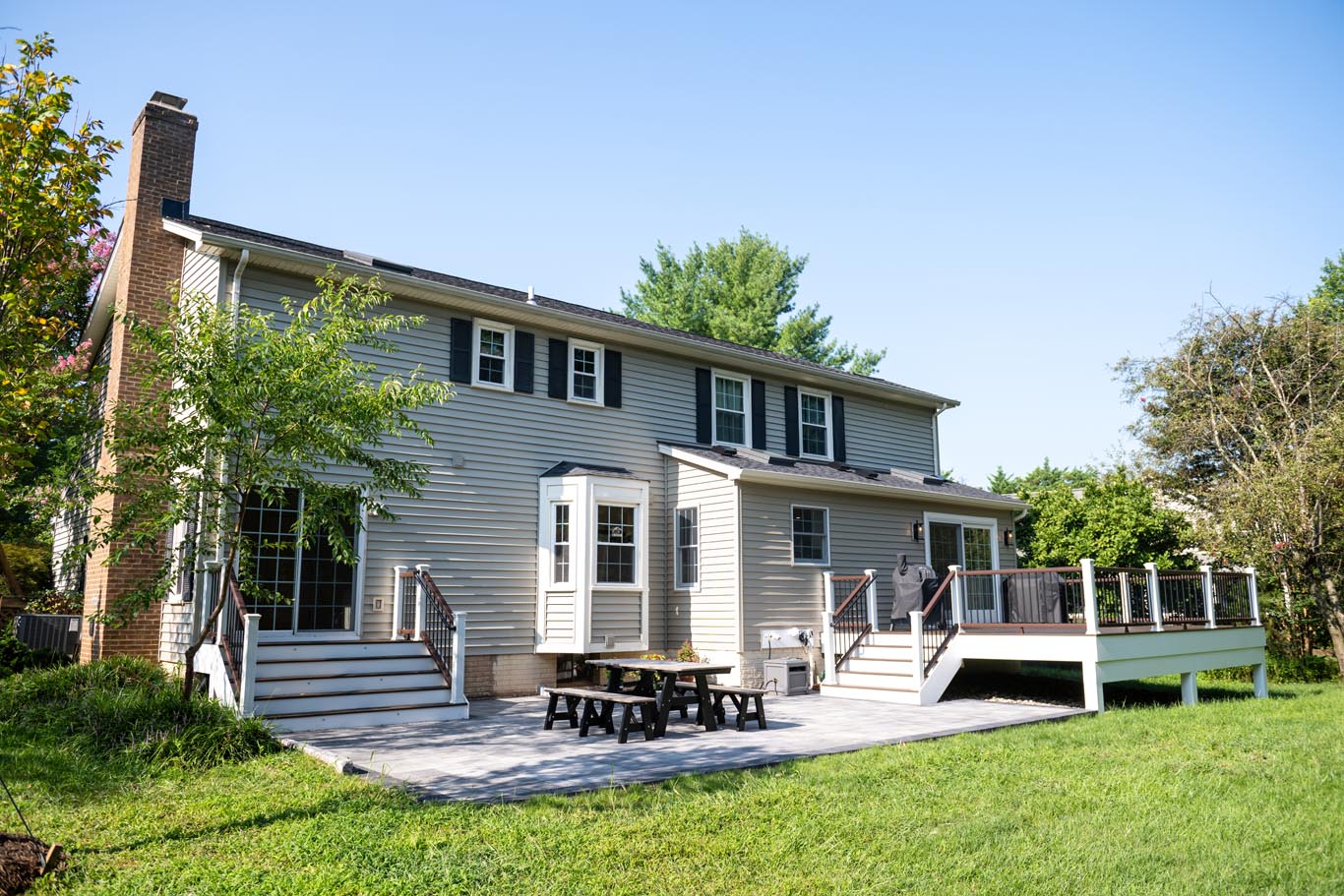 Image by Lesnick Photo Two-story house with a large backyard deck featuring a picnic table, perfect for home additions in Northern Virginia. The light-colored exterior boasts multiple windows and a chimney on the left, while the grassy yard is dotted with trees, creating an inviting outdoor space.