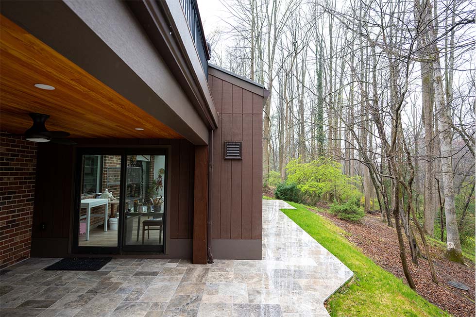 NorthwoodConstruction-Home-additions-northern-virginia-wood ceiling The patio area of this modern house, featuring a wooden ceiling and stone flooring, is a stunning result of home additions in Northern Virginia. Its glass door opens to a serene wooded area with bare trees and hints of greenery, perfect for embracing nature's tranquility.