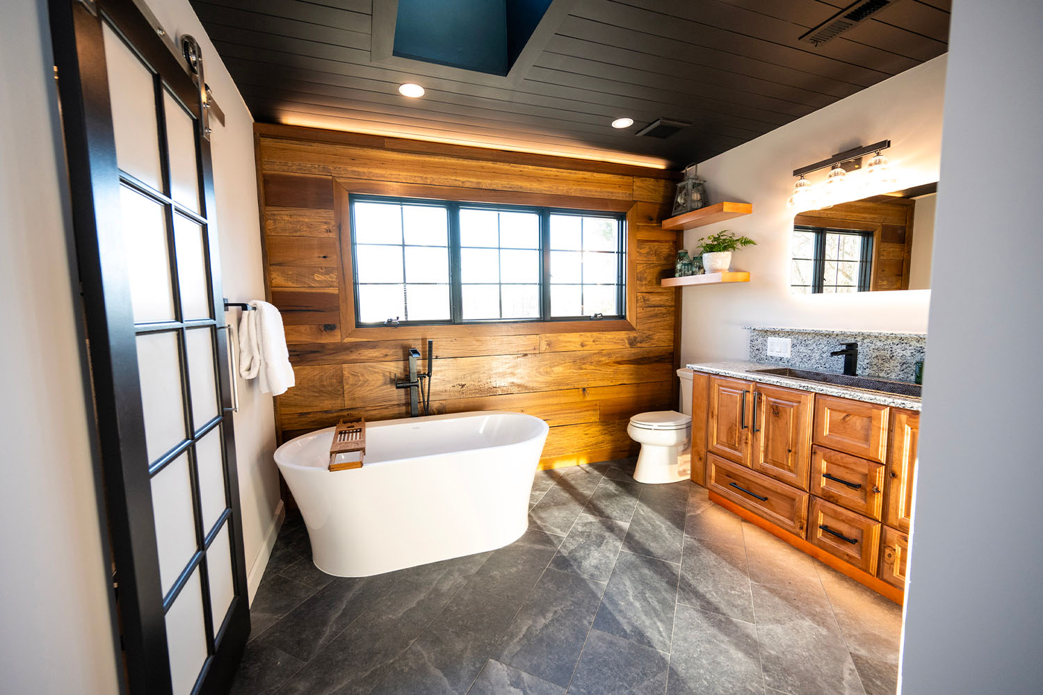 Explore bathroom remodeling in Northern Virginia with a modern touch: a freestanding bathtub, wooden walls, black framed window, and sleek cabinets create an inviting retreat.