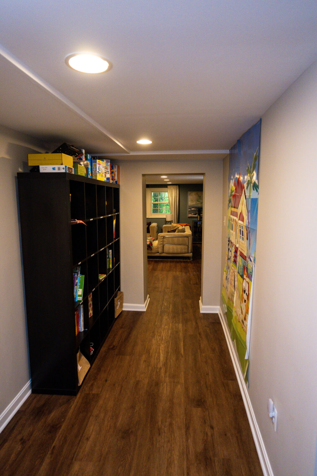 Image by Lesnick Photo A narrow hallway with wooden flooring features a black shelf holding board games on the left and a colorful mural on the right. The hallway leads to a room with a sofa and window.