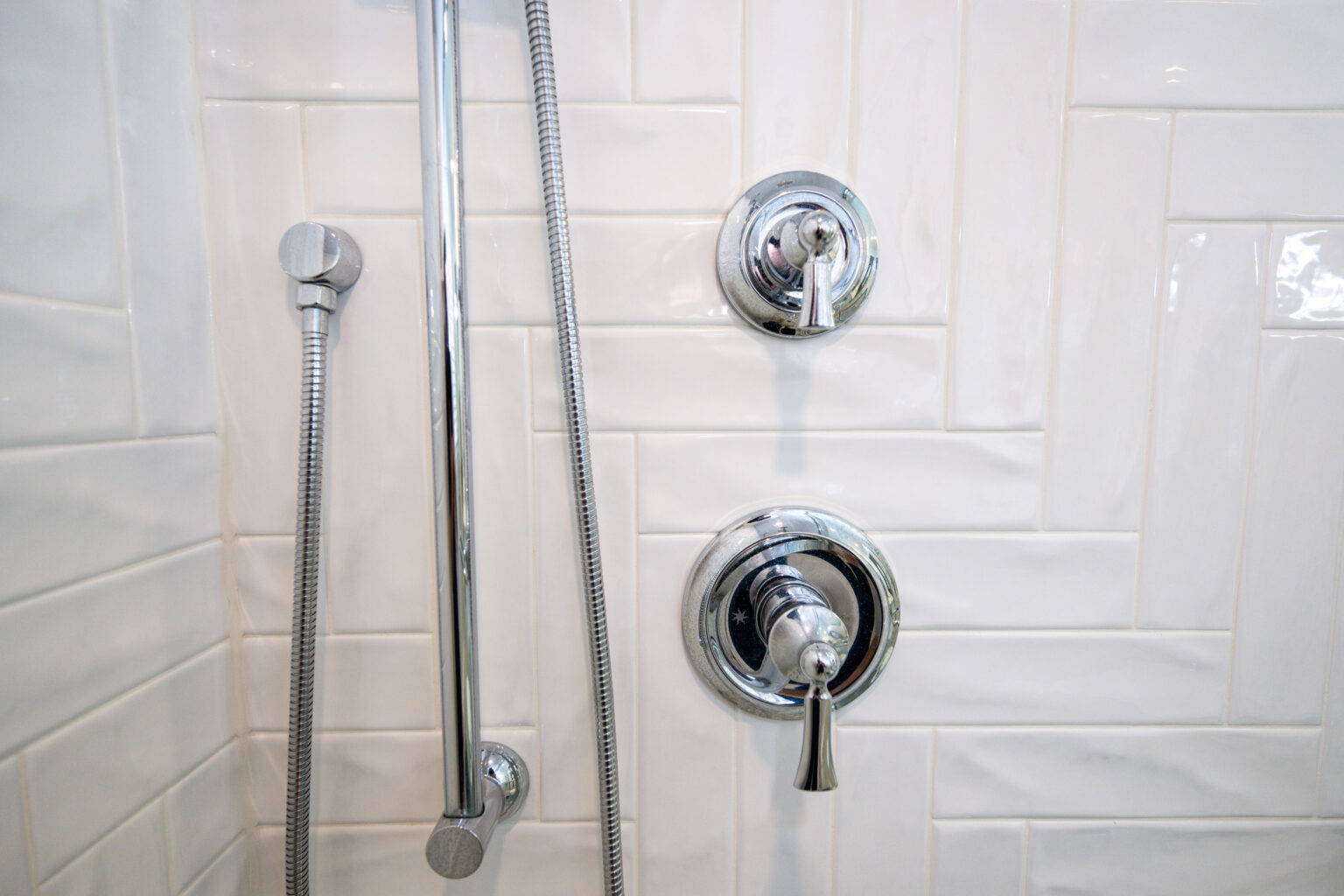 Image by Lesnick Photo Close-up of a shower wall featuring two chrome controls and a vertical handheld showerhead with a hose against white subway tiles.