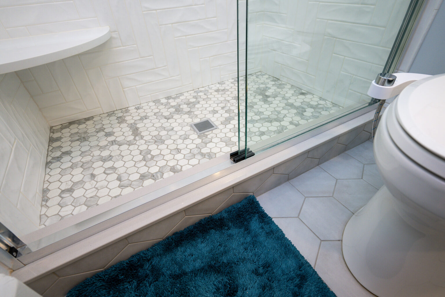 Image by Lesnick Photo Bathroom with a hexagonal tiled shower floor, white tiled walls, clear glass door, white toilet, and a blue textured bath mat.