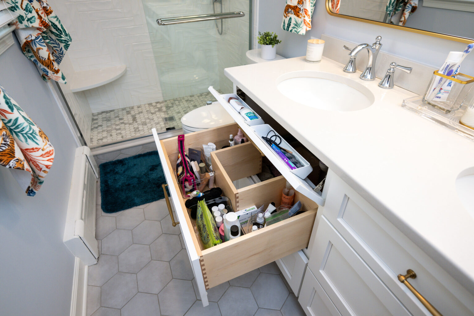 Image by Lesnick Photo Bathroom with a white vanity and open drawers displaying toiletries. A walk-in shower with a glass door is visible. The floor has hexagonal tiles, and floral towels hang nearby.