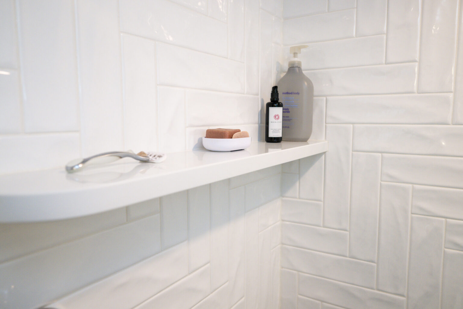 Image by Lesnick Photo A white shower shelf holds a spoon, soap dish with soap, a small bottle, and a large pump bottle, all against white tiled walls.