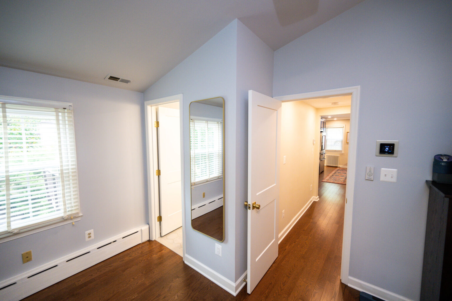 Image by Lesnick Photo Small room with white walls, hardwood floor, and a window with blinds. A tall mirror leans against the wall near an open door leading to a hallway. A bathroom is visible on the left.