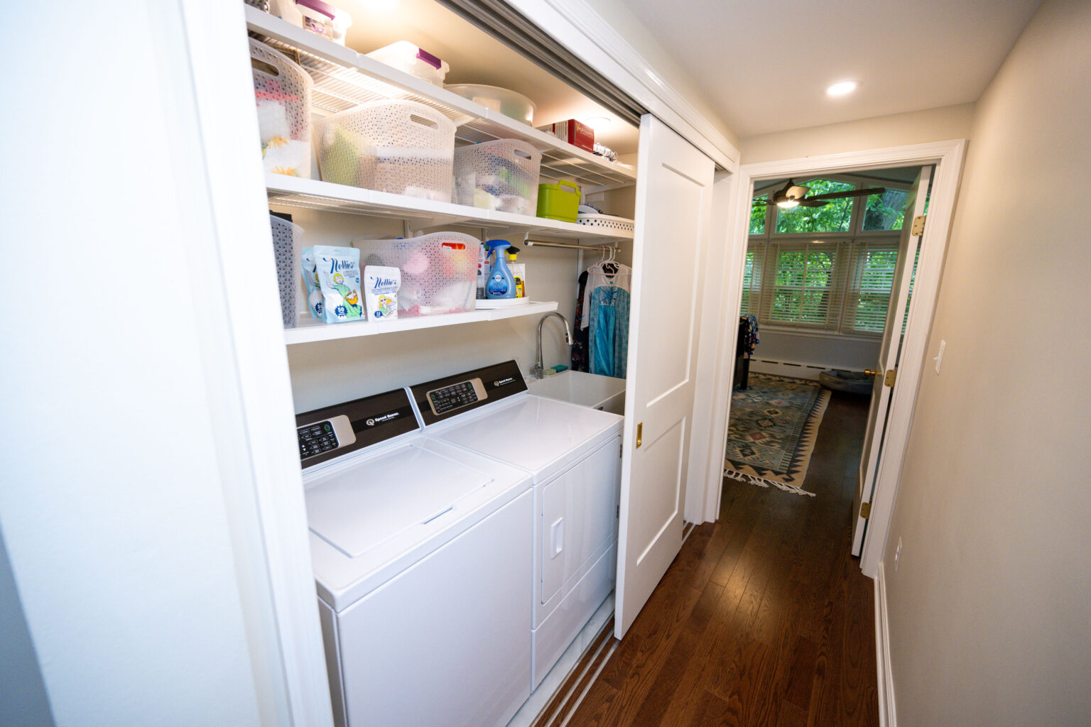 Image by Lesnick Photo Compact laundry area with washer, dryer, and shelves stocked with cleaning supplies. Sliding doors open to a wood-floored hallway leading to a well-lit room with large windows.