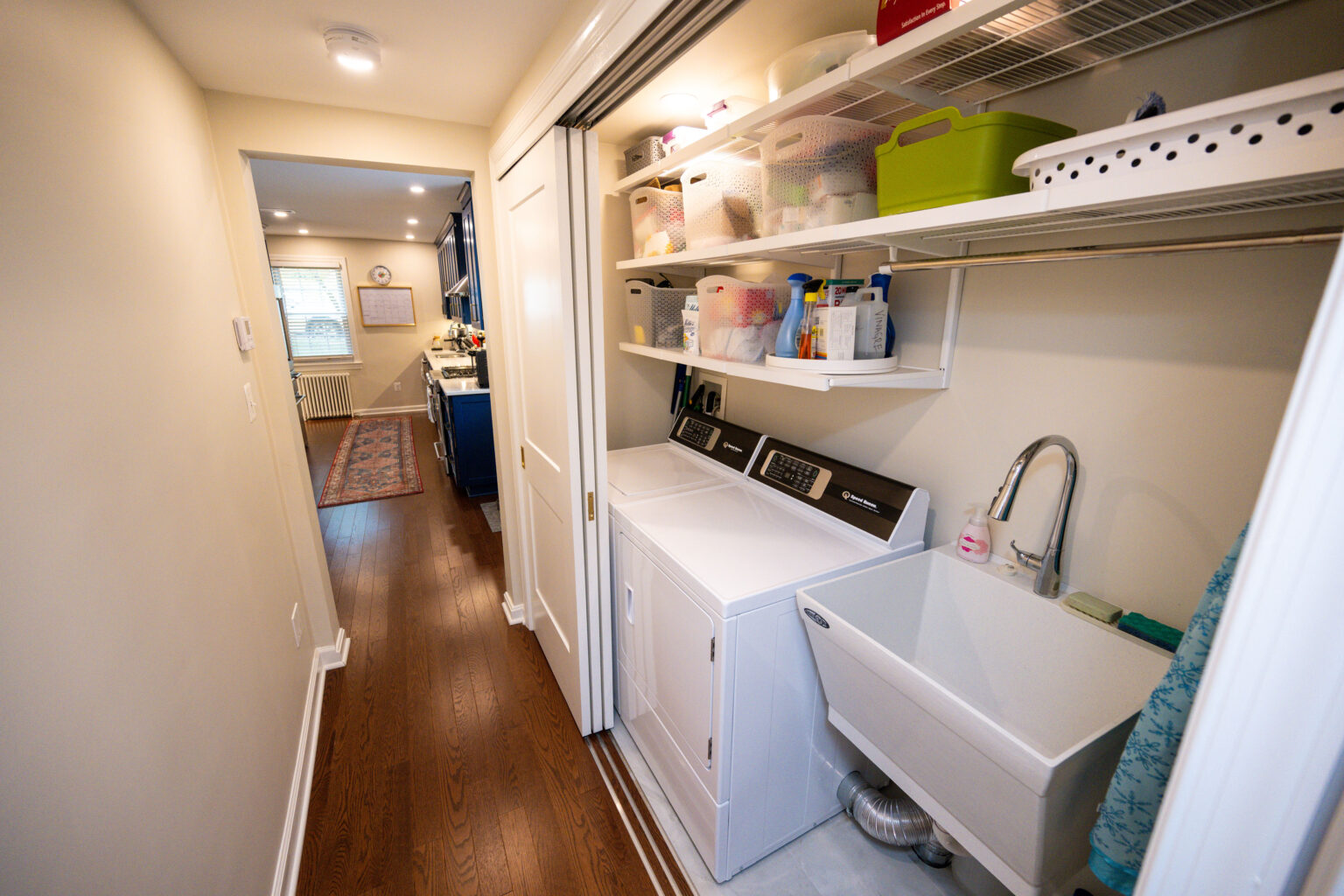 Image by Lesnick Photo Laundry room with a washing machine, sink, shelves with storage bins, and cleaning supplies. The room has a wooden floor and leads to a well-lit hallway.