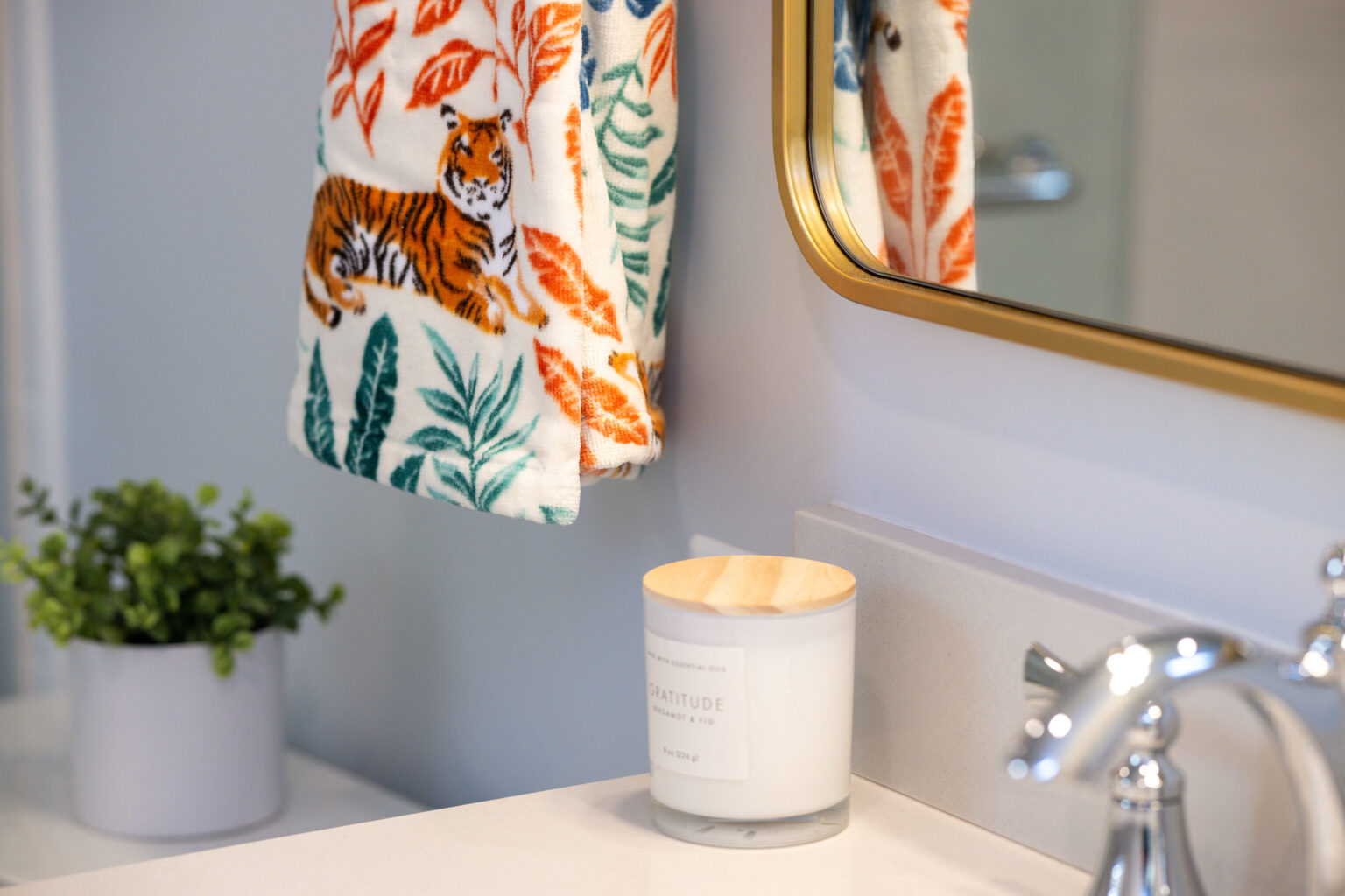 Image by Lesnick Photo Bathroom scene with a tiger-patterned towel, gold-framed mirror, potted plant, and a candle labeled 'Gratitude' on a white countertop.