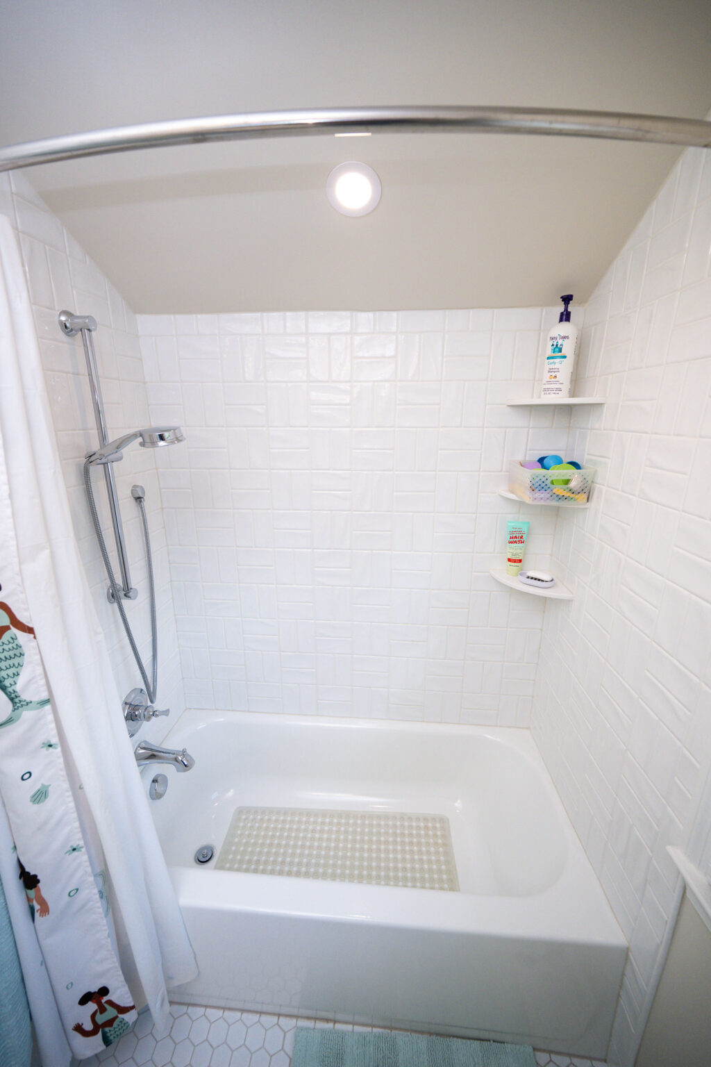 Image by Lesnick Photo White tiled bathroom with a bathtub and a shower head. Shelves hold toiletries and a sponge. The floor has hexagonal tiles, and the ceiling light is on. A shower curtain features a green pattern.
