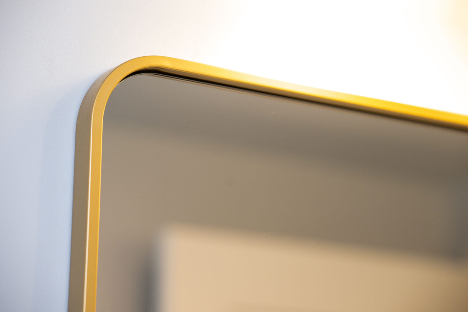 Image by Lesnick Photo Close-up of the corner of a rectangular mirror with a thin gold frame, mounted on a light-colored wall.
