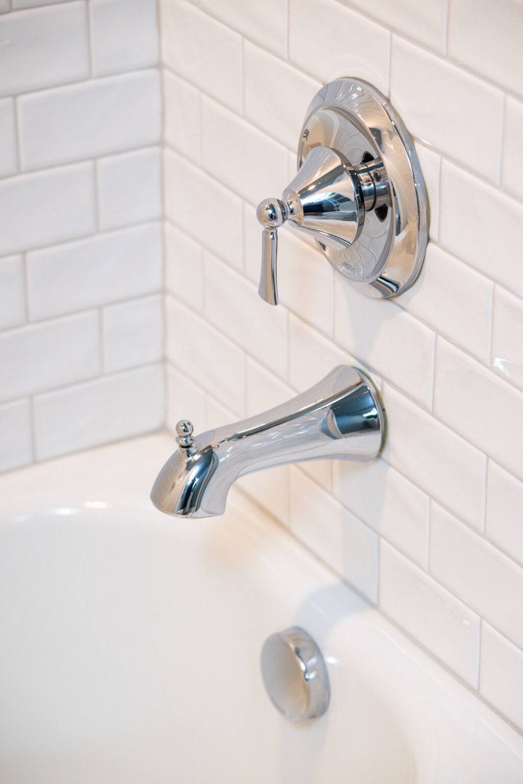 Image by Lesnick Photo Chrome bathtub faucet and spout set against a white tiled wall, with a drain stopper visible below.