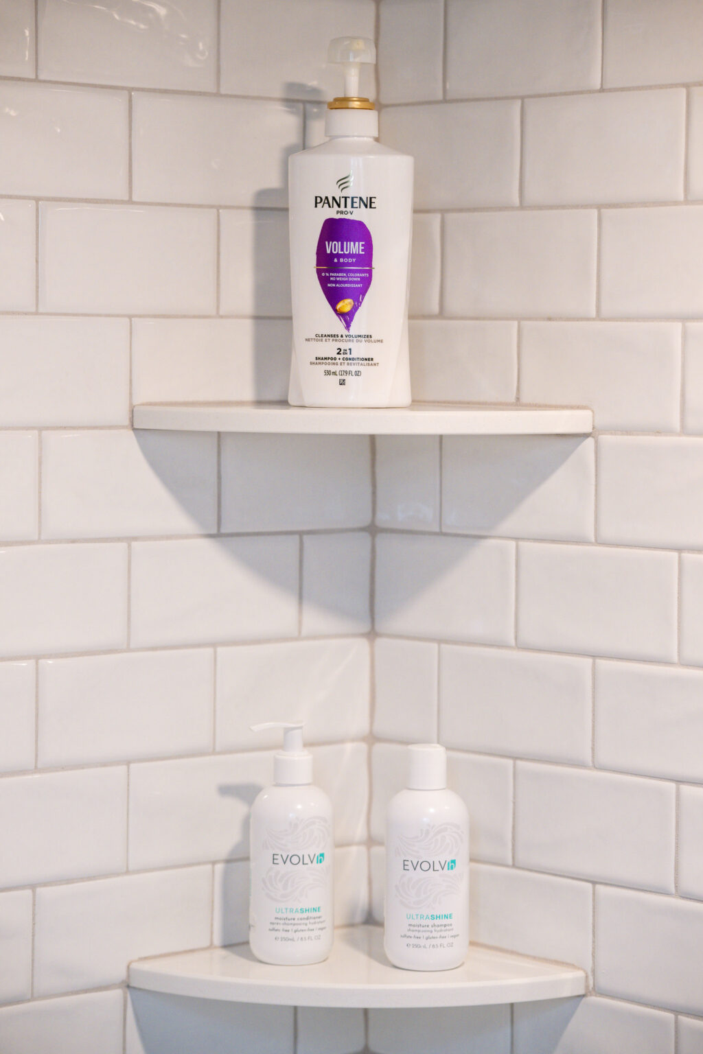 Image by Lesnick Photo Shower shelves with Pantene volume shampoo on top shelf and Evolvh hair products on bottom shelf, set against white subway tiles.