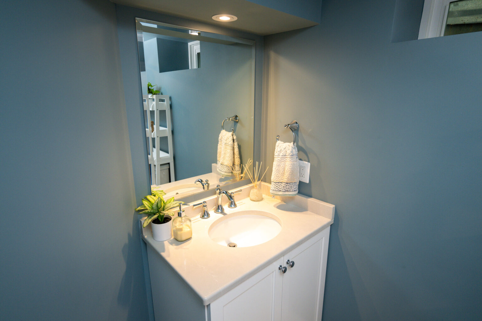 Image by Lesnick Photo A small bathroom with a white vanity and sink, a mirror, towels, a potted plant, and toiletries. The walls are painted blue, and there is overhead lighting.