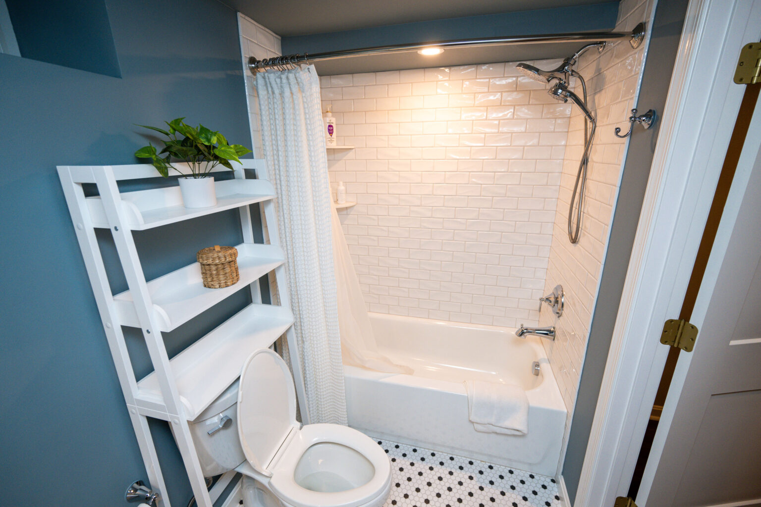 Image by Lesnick Photo Bathroom with a white tiled bathtub and shower, a white toilet, a shelving unit with a plant and basket, and a black and white patterned floor.