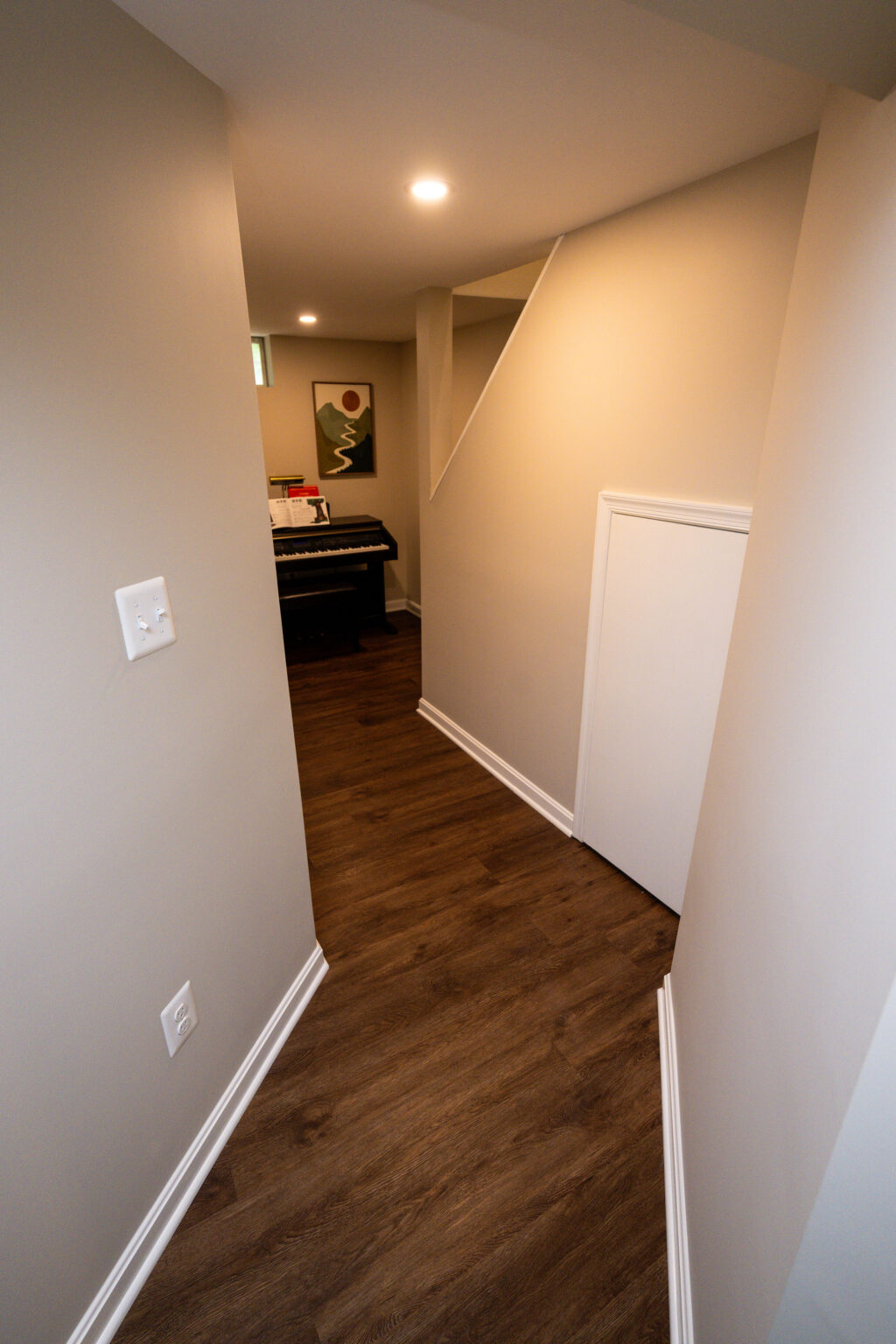 Image by Lesnick Photo Hallway with beige walls leading to a room with a piano under a small window. A piece of abstract art is visible on the back wall. The floor is wood with white baseboards.