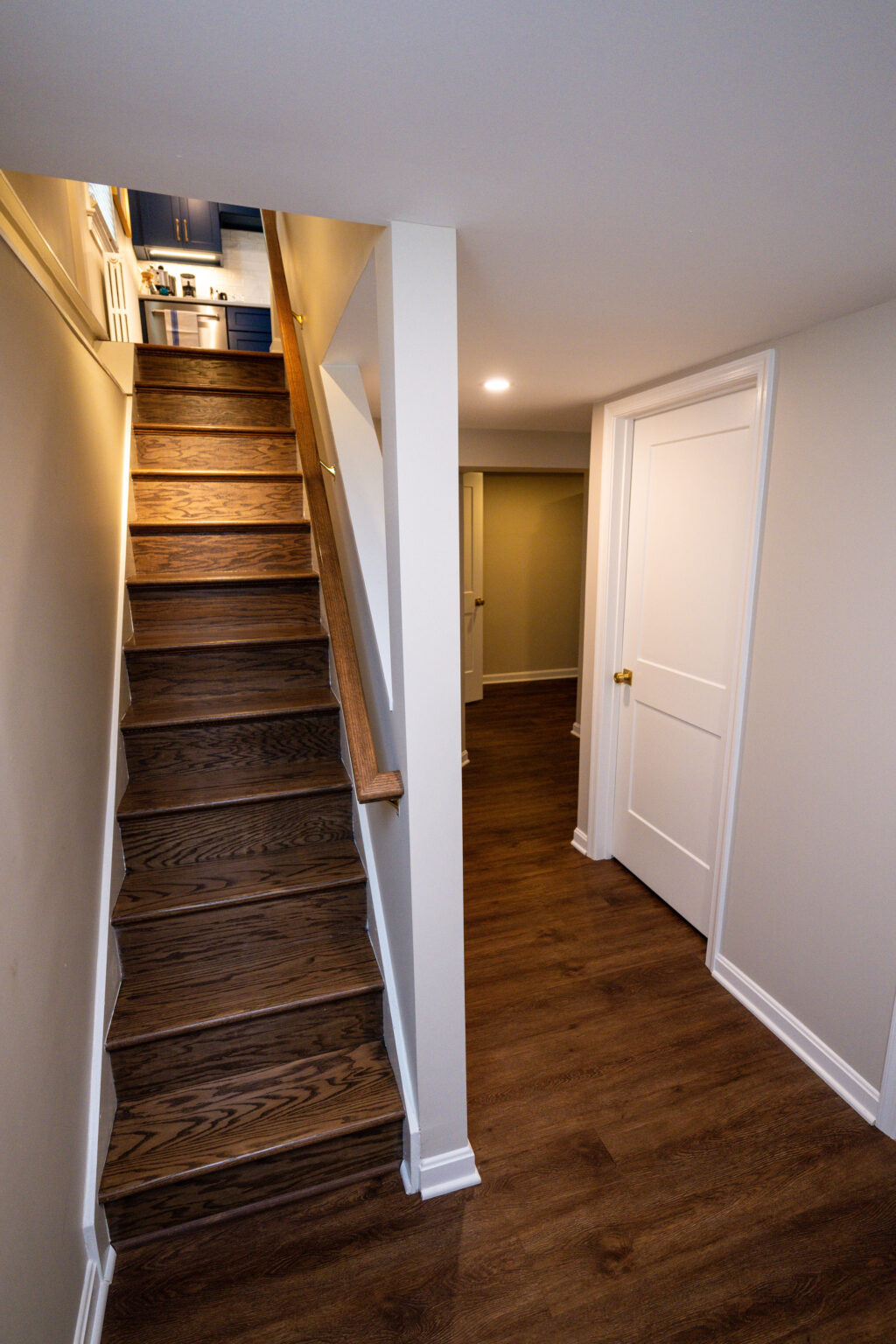 Image by Lesnick Photo A wooden staircase leads upwards beside a hallway with closed white doors on a brown floor.