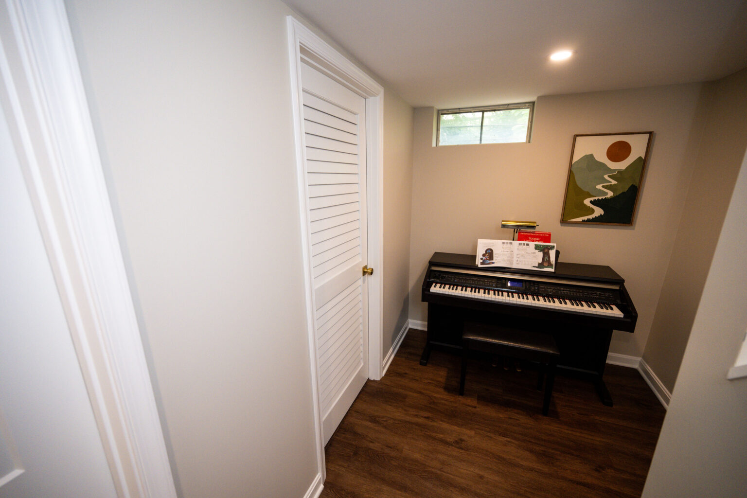 Image by Lesnick Photo Small room with electronic piano, sheet music, and a bench. Minimalist decor features a landscape painting, small window, and wood flooring. Door with louvered closet is visible.
