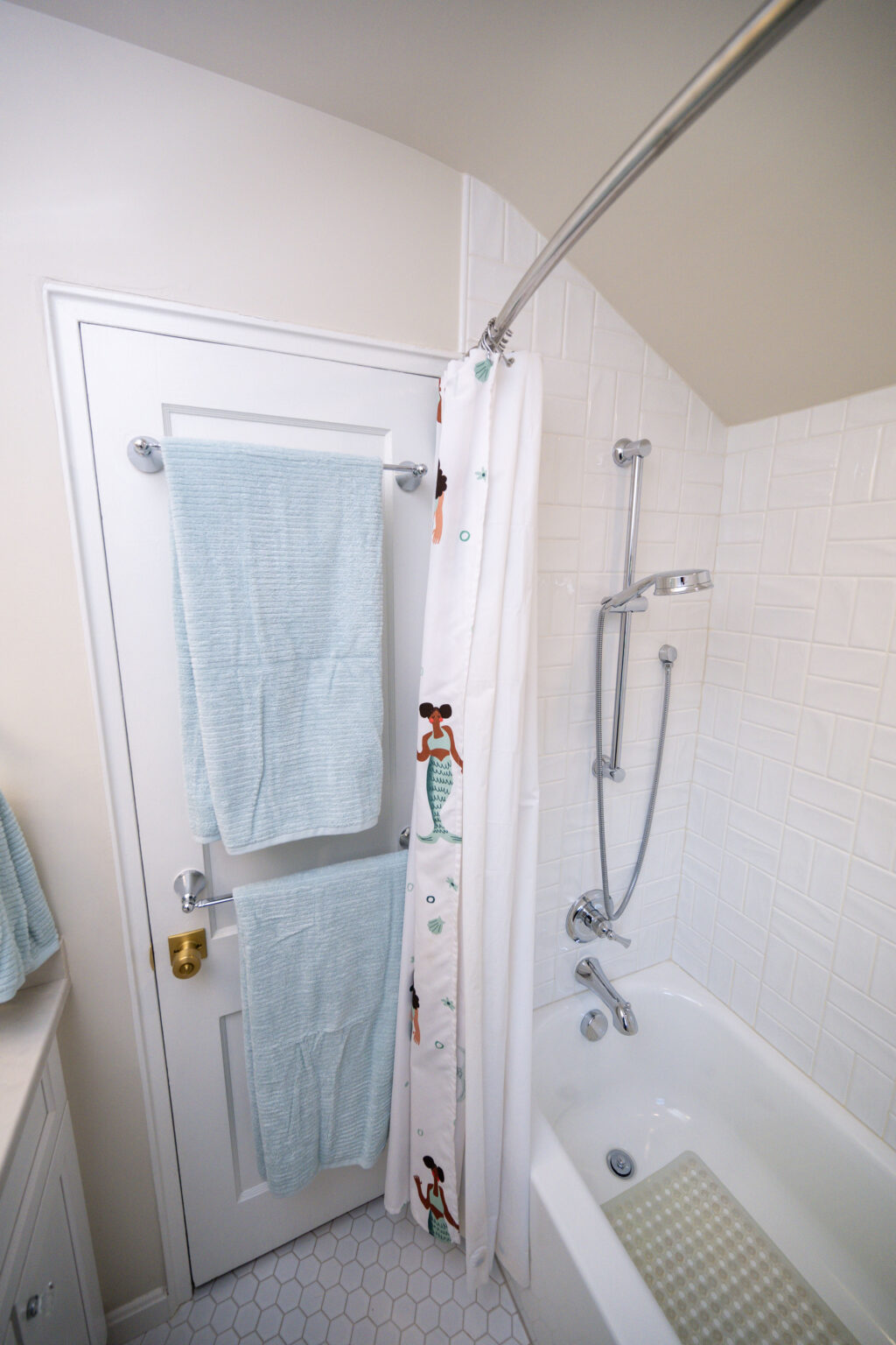 Image by Lesnick Photo Bathroom with a bathtub, showerhead, curved curtain rod, and a closed door. Blue towels hang on the door; the shower curtain features cartoon prints. White tile walls and hexagonal floor tiles.