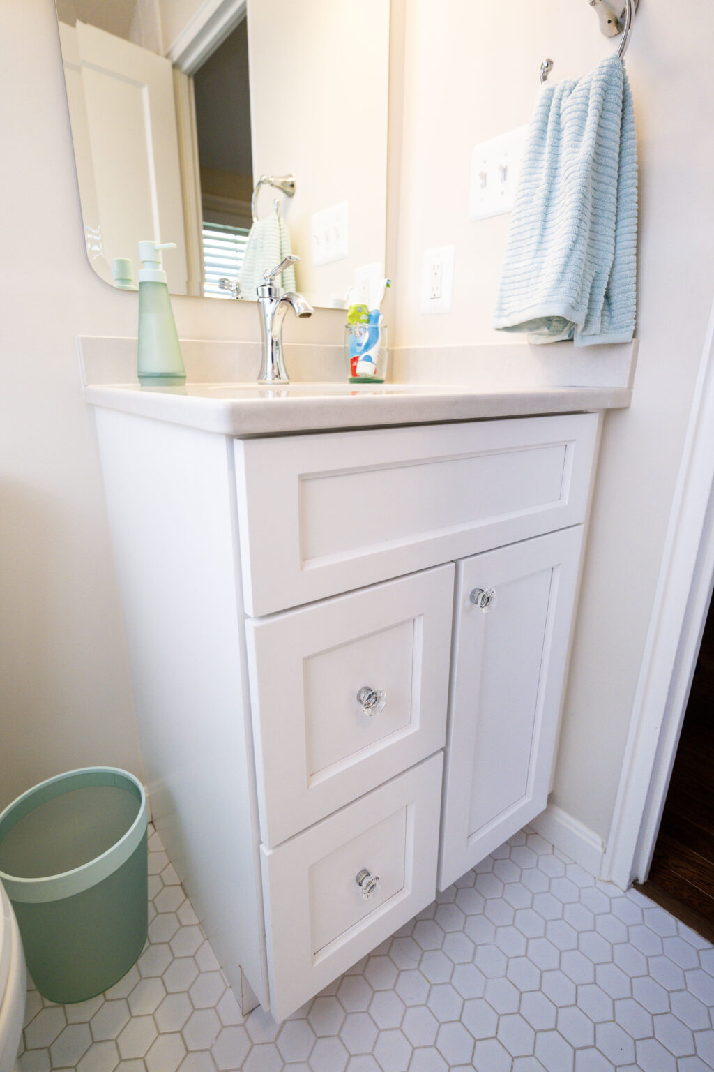 Image by Lesnick Photo White bathroom vanity with three drawers, a sink, liquid soap dispenser, and hand towel. A mirror is above the countertop. A blue waste bin is on the hexagonal-tiled floor.