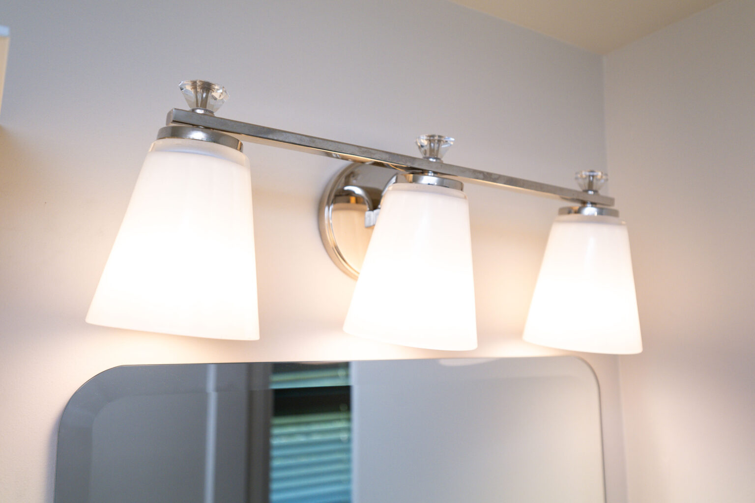 Image by Lesnick Photo Wall-mounted light fixture with three conical white lampshades above a mirror.