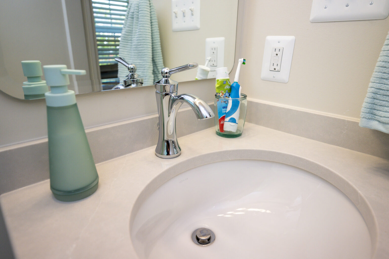 Image by Lesnick Photo Bathroom sink with a chrome faucet, liquid soap dispenser, and a cup holding toothbrushes and toothpaste. A towel hangs on the right near a power outlet.