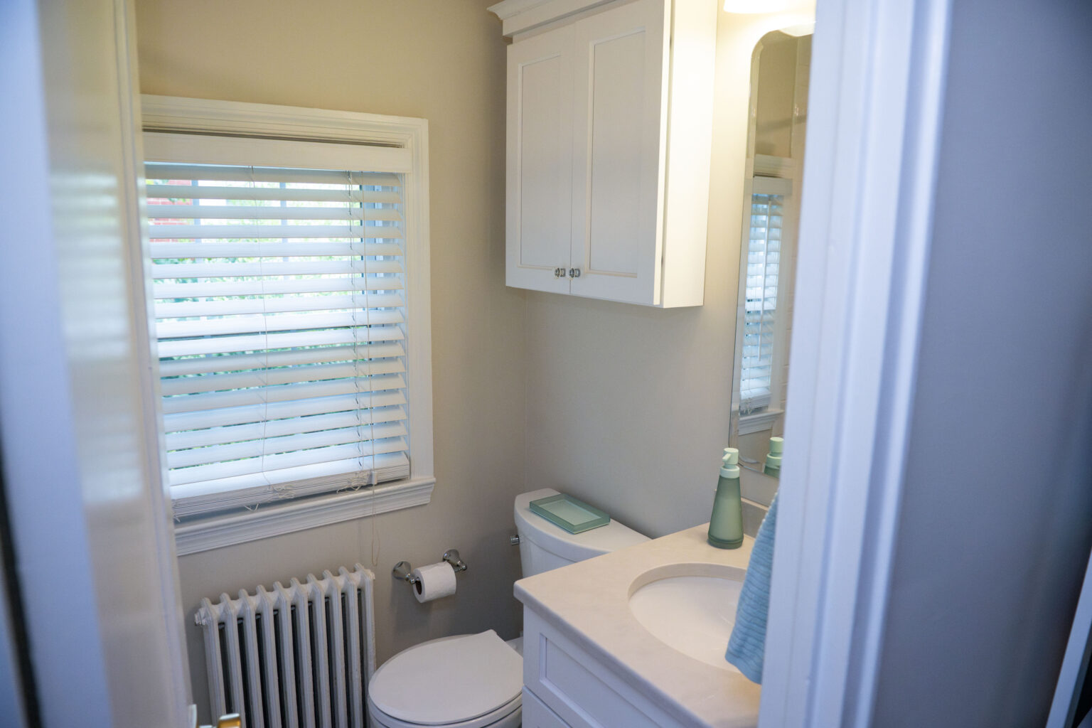 Image by Lesnick Photo A small bathroom with a toilet, sink, and mirror. It has a window with blinds, a radiator, and white cabinets above the toilet.