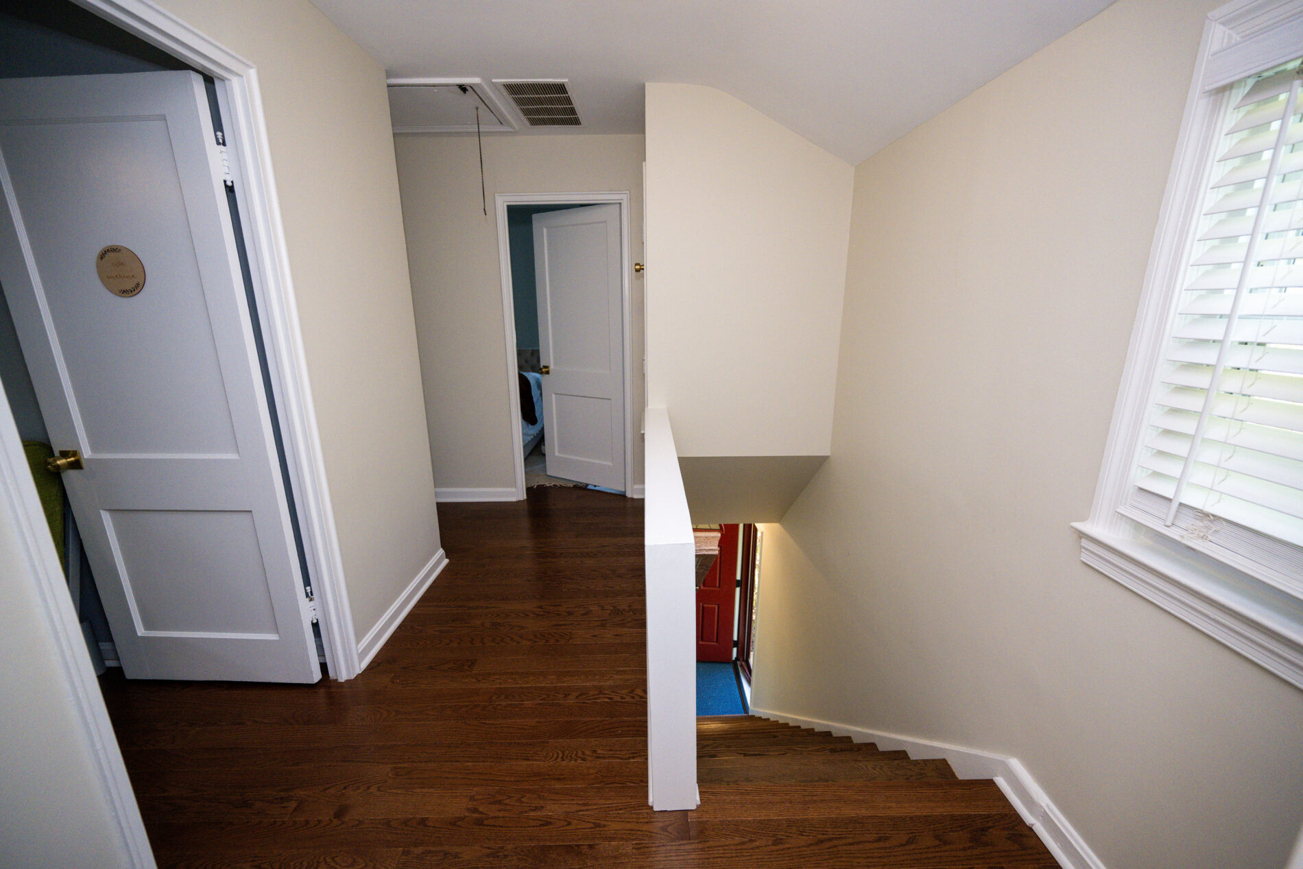 A hallway with wooden flooring leads to a staircase going down. Two white doors are on the left, and a window with blinds is on the right.