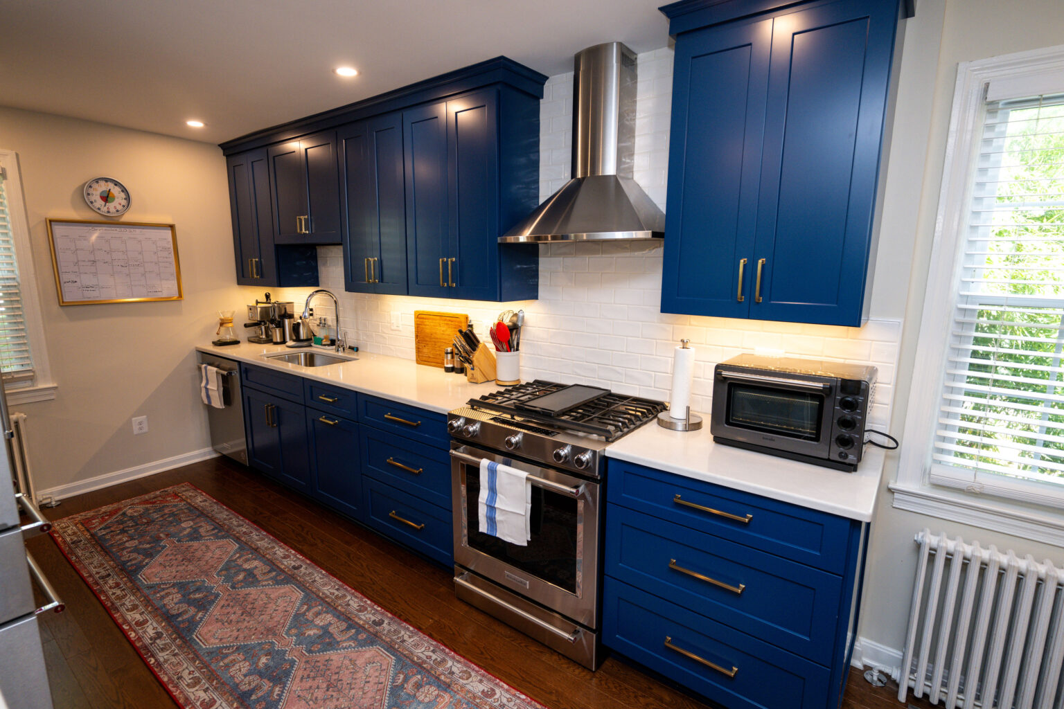 Image by Lesnick Photo A kitchen with blue cabinets, a stainless steel stove, and a range hood. White countertops and a red patterned rug complete the decor.