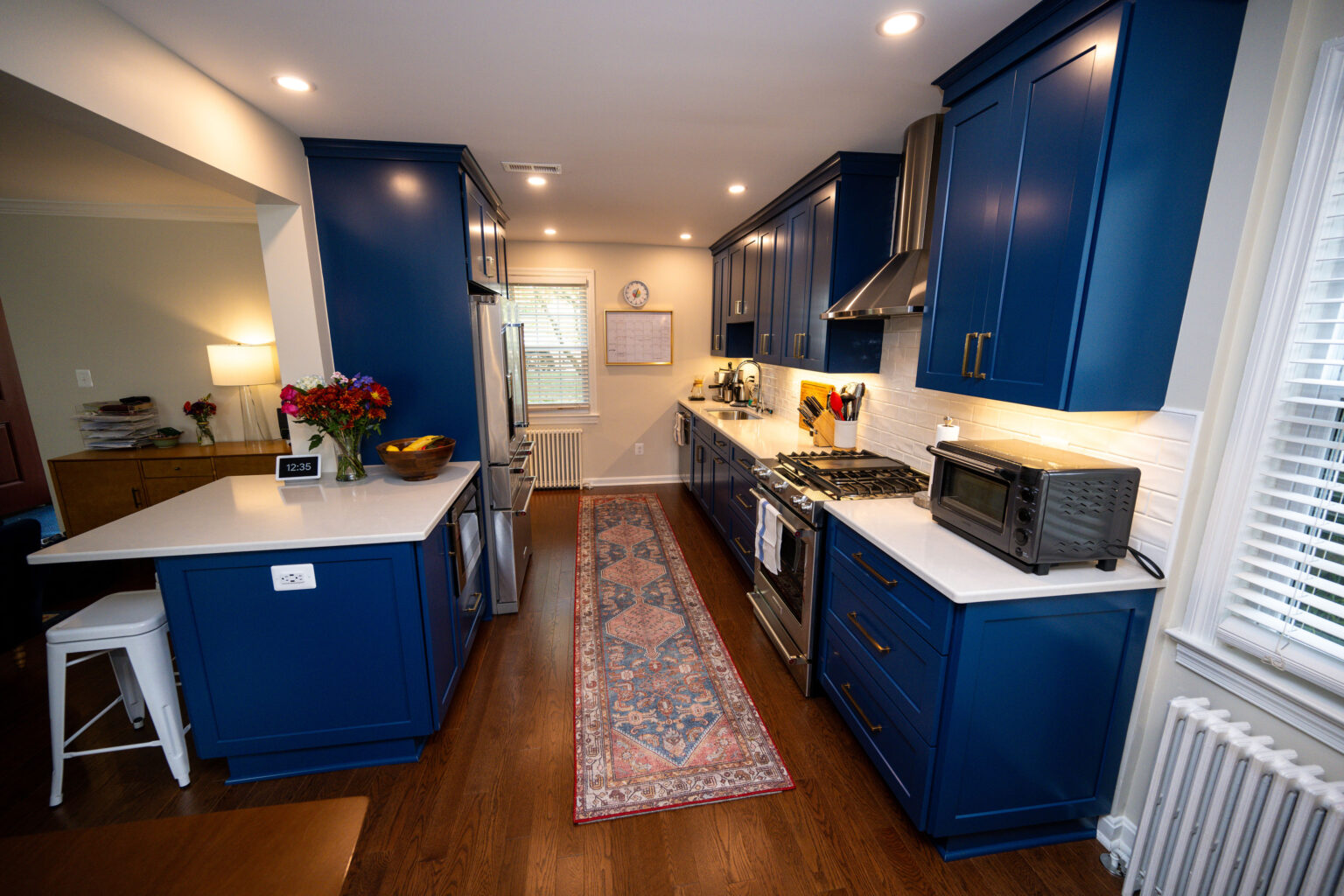 Image by Lesnick Photo A modern kitchen featuring blue cabinets, white countertops, a stainless steel oven and hood, and a long patterned rug on wooden flooring. There are flowers and a fruit bowl on the counter.
