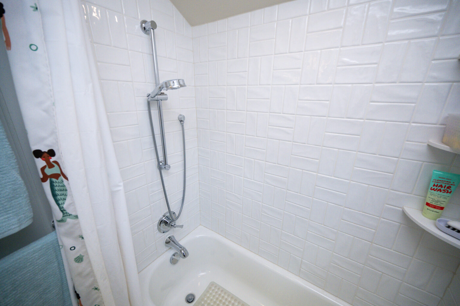 Image by Lesnick Photo White-tiled bathtub with a showerhead and faucet, a toiletry bottle on a corner shelf, a white shower curtain with a pattern on the left.