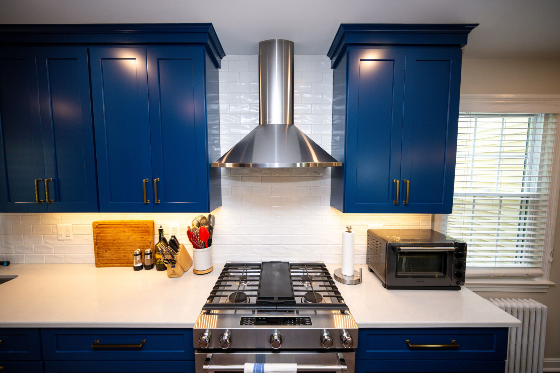 Modern kitchen with blue cabinets, stainless steel range hood, white subway tile backsplash, gas stove, toaster oven, and window with blinds.