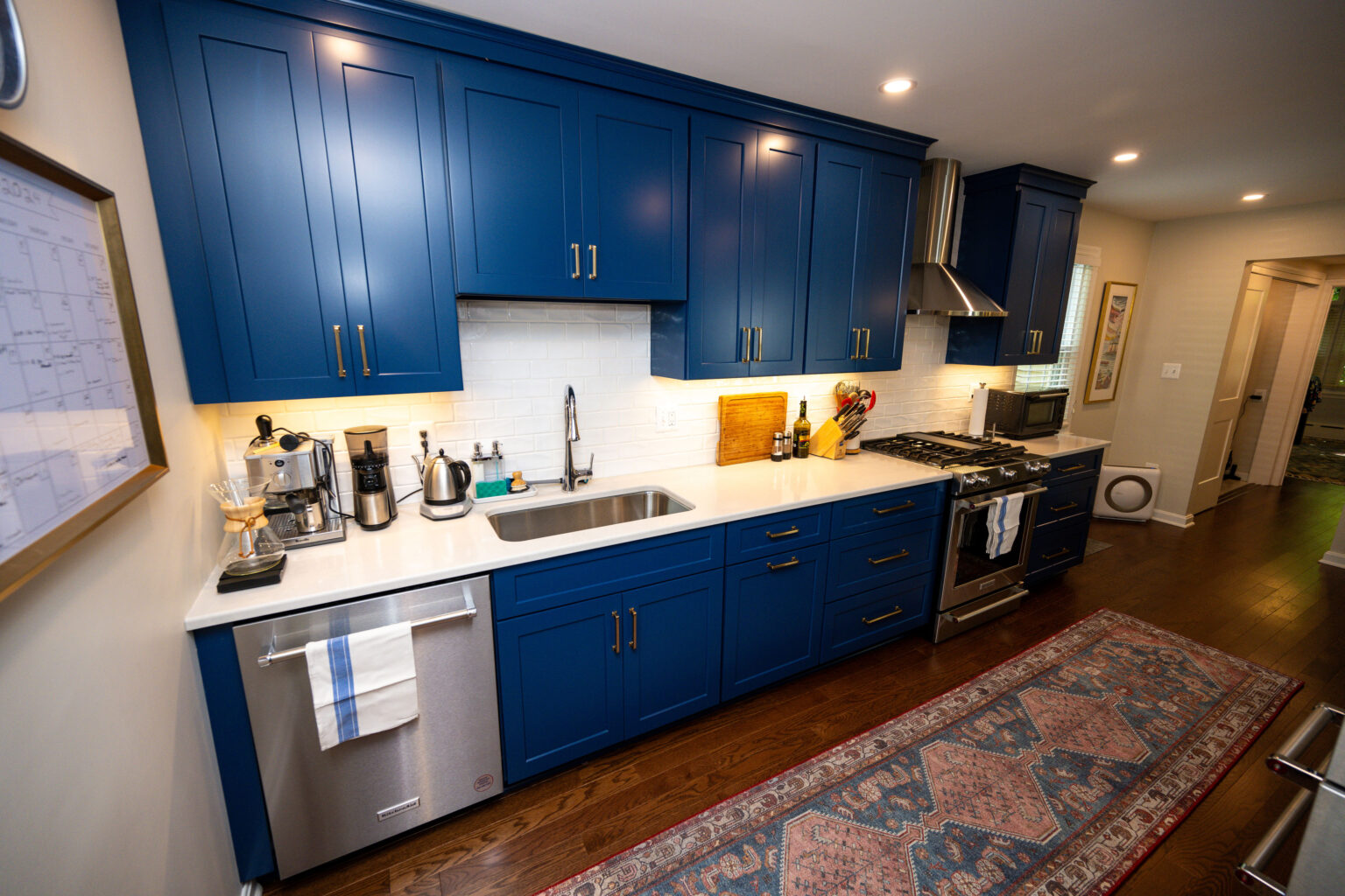Image by Lesnick Photo Modern kitchen with blue cabinets, white countertops, stainless steel appliances, and a patterned rug. Bright lighting and wood flooring complete the space.