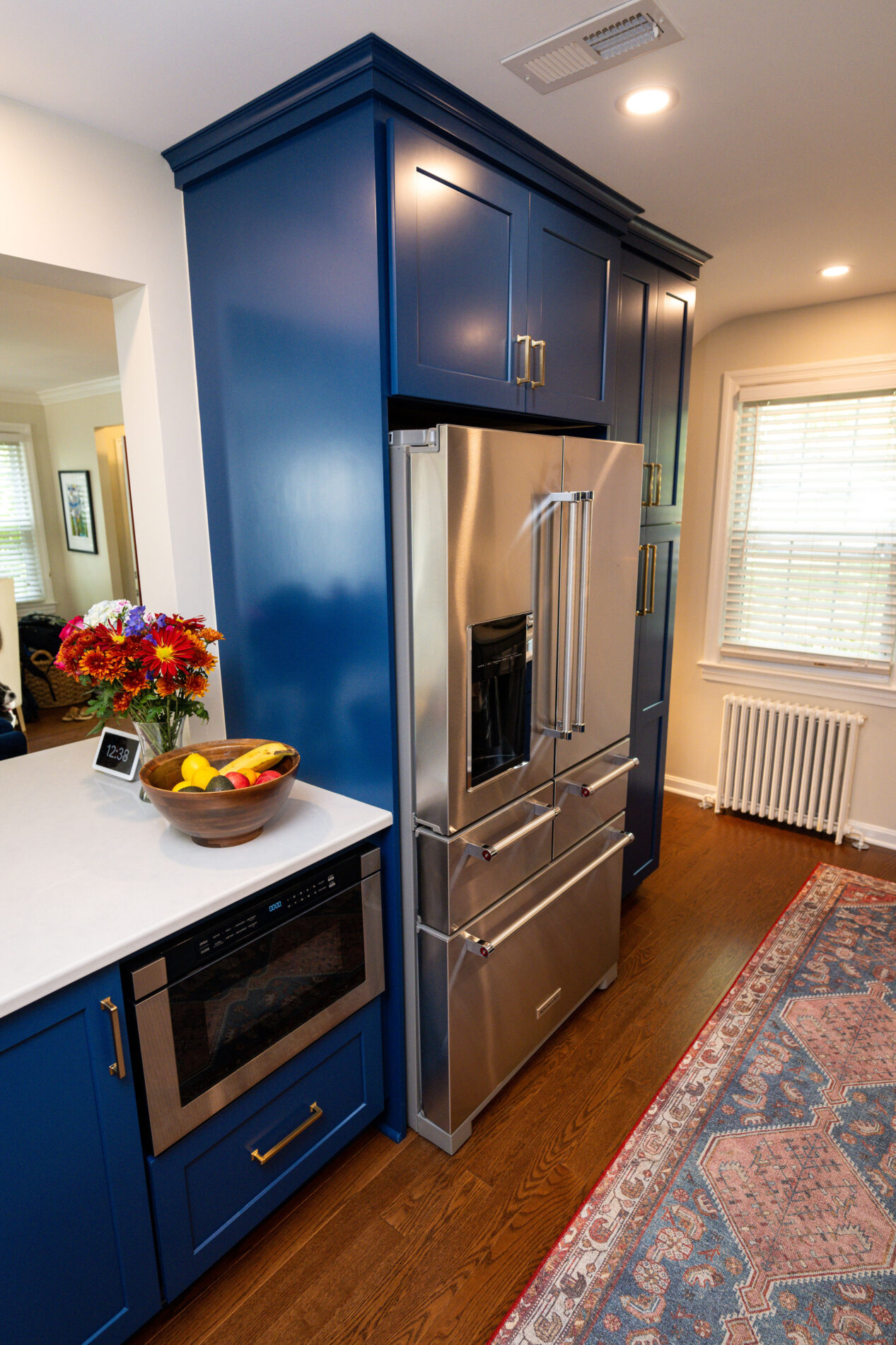 A modern kitchen with blue cabinets, a stainless steel refrigerator, built-in oven, and a white countertop featuring a bowl of fruit and a vase with flowers. A patterned rug is on the wooden floor.