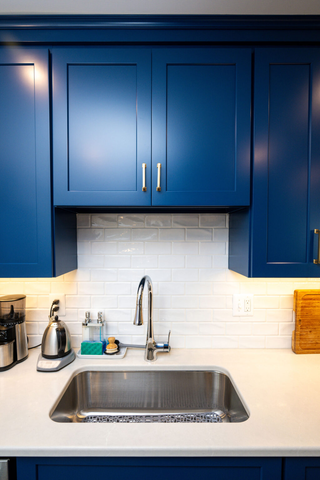 Image by Lesnick Photo Modern kitchen with blue cabinets, a stainless steel sink, and white subway tile backsplash. There is a kettle, soap, and a wooden cutting board on the countertop.