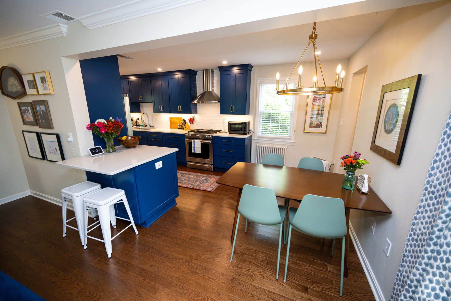 Modern kitchen and dining area with blue cabinets, white countertops, a wooden table with green chairs, and framed art on walls. Chandelier above the table and a patterned rug on the wooden floor.