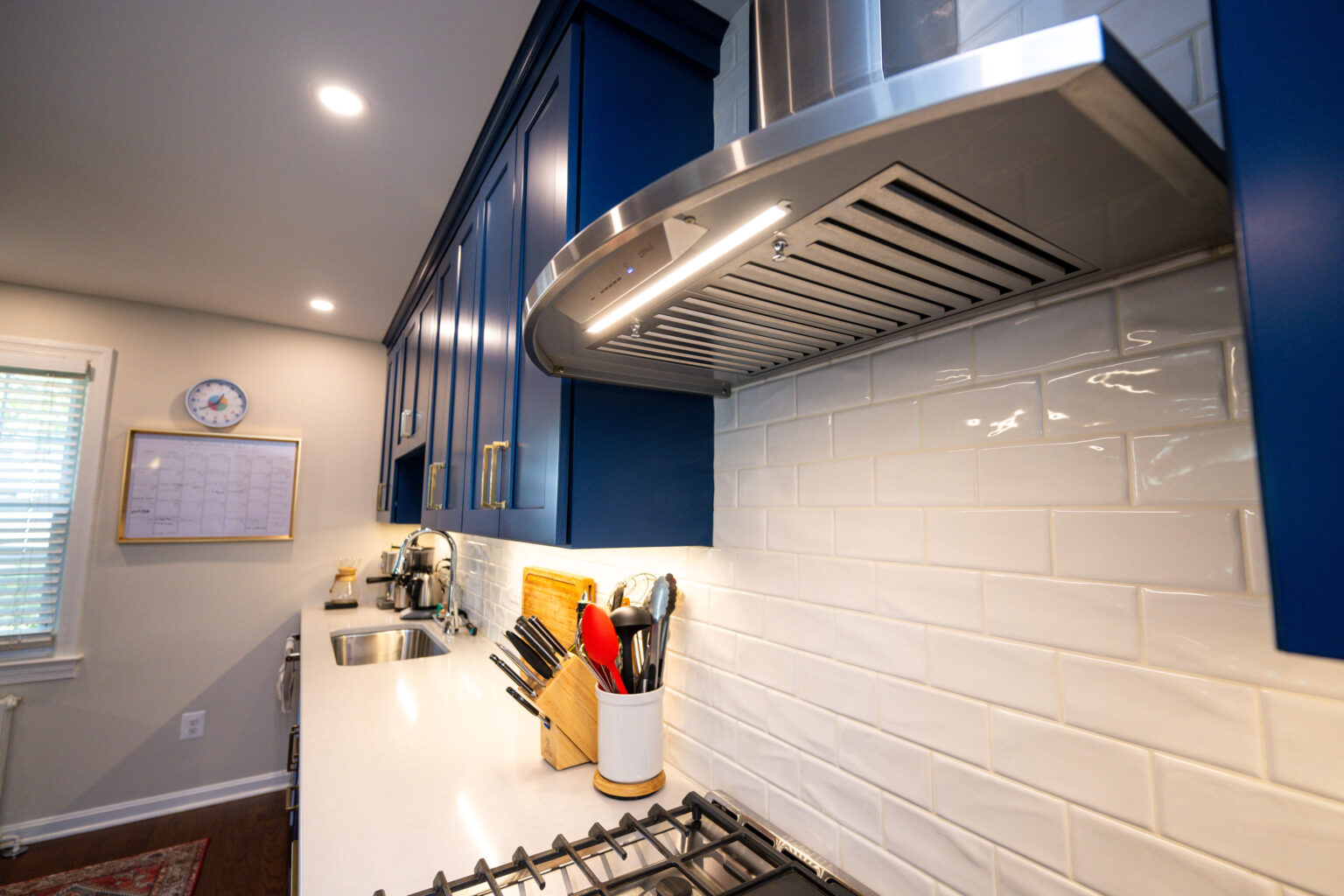 Image by Lesnick Photo Modern kitchen with blue cabinets, white subway tile backsplash, and stainless steel range hood. Countertop features a sink, knife block, and utensils. Wall-mounted clock and calendar visible.