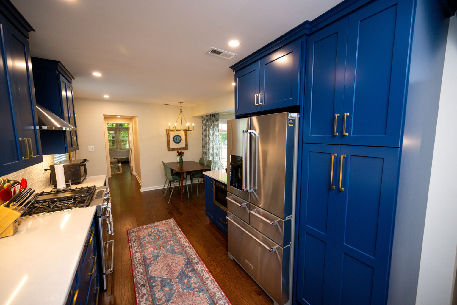Image by Lesnick Photo A modern kitchen with blue cabinets, stainless steel appliances, and a decorative rug. A dining area with a hanging light fixture is visible in the background.