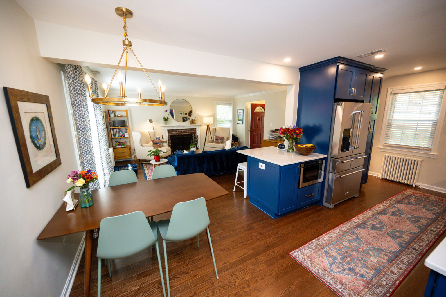 Image by Lesnick Photo Open-concept kitchen and dining area with blue cabinetry, wooden dining table with green chairs, modern chandelier, and adjoining living room with sofas and a fireplace.