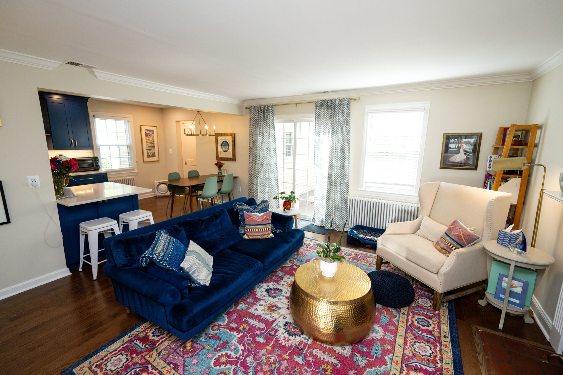 Living room with a blue sofa, colorful rug, gold coffee table, and armchair. Dining area with blue chairs and a table is in the background. Large windows bring in natural light.